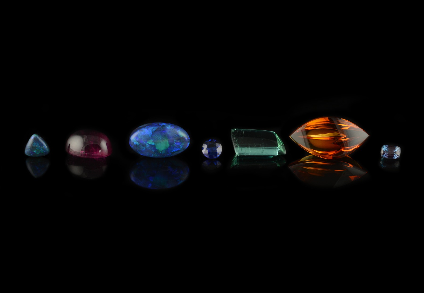 Loose gemstones for commissions, from left to right: black opal, rubellite tourmaline, White cliffs black opal, sapphire, petrol blue trapezoid tourmaline, citrine, alexandrite